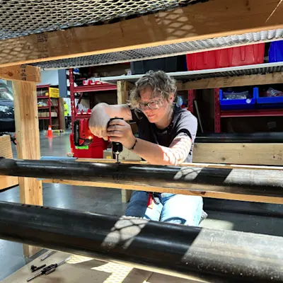 Technician wearing safety glasses uses a power drill on a metal pipe in a warehouse setting.