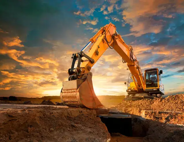 Yellow excavator digging a trench at a construction site against a sunset sky.