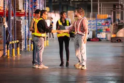 Warehouse workers in safety vests shaking hands during a meeting in a large distribution center.