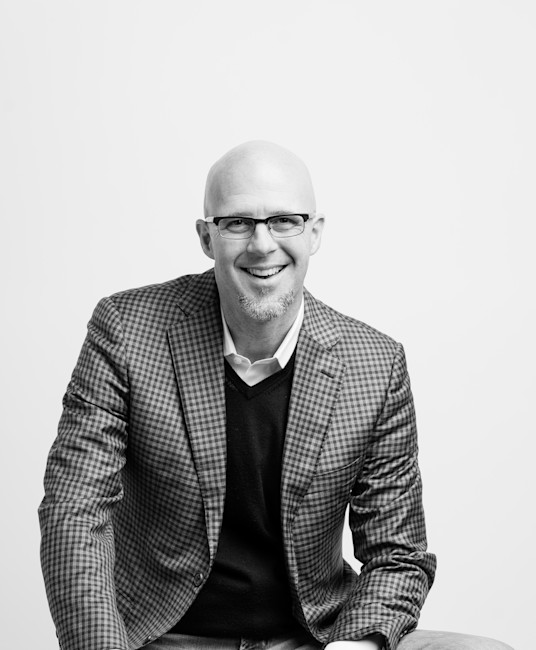 Black-and-white portrait of smiling bald man with glasses and goatee in plaid blazer seated
