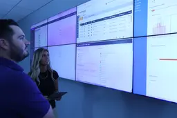 Two colleagues looking at a large wall of digital data monitors in an operations center.