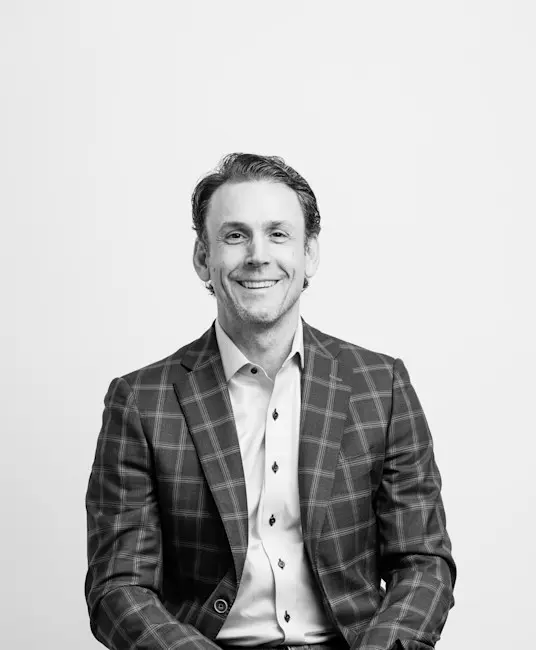 Black-and-white portrait of smiling man in plaid blazer and button-down shirt seated against plain background