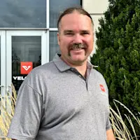 Smiling man with a mustache, Luke N., wearing a grey polo shirt in front of a building.