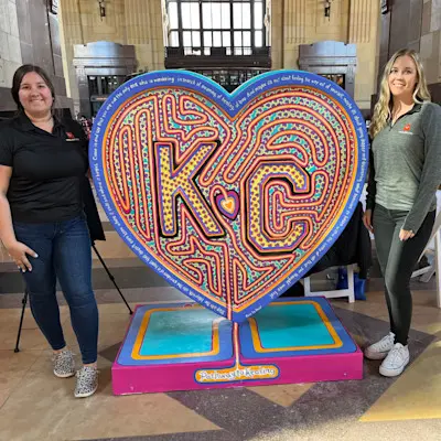 Two women posing next to a large, colorful heart sculpture with "KC" labyrinth art.