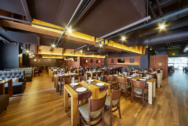 Modern restaurant interior with wooden tables, set place settings, and industrial-style ceiling lighting.