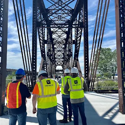 A group of Velociti engineers wearing safety vests and hard hats walking across a large steel truss bridge.