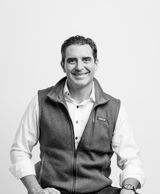 Black-and-white portrait of smiling man seated, wearing fleece vest over button-down shirt