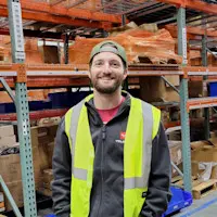Smiling man, Ryan Blum, wearing a backwards baseball cap and a neon safety vest in a warehouse setting.