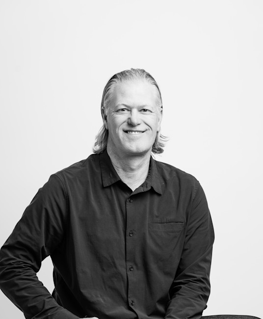 Black-and-white portrait of smiling man with shoulder-length hair wearing a dark button-up shirt