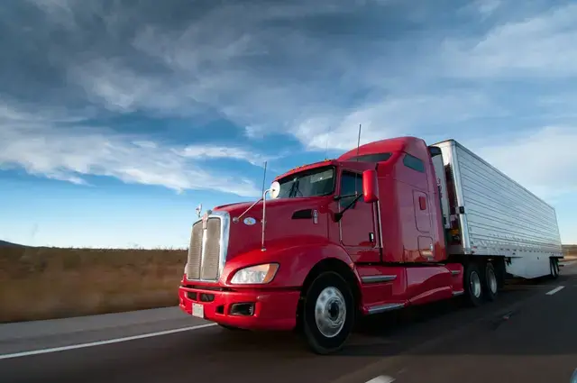 A bright red semi-truck hauling a white trailer on a highway under a blue sky.
