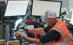 A technician wearing an orange safety vest and a grey cap using a handheld scanner to process equipment at a workstation with multiple monitors.