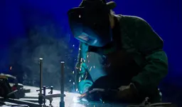 A welder wearing a protective mask and green jacket working on a metal project with bright sparks flying.