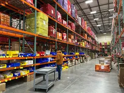 A worker standing next to a metal cart in a large warehouse aisle with tall shelving units filled with colorful bins and packages.