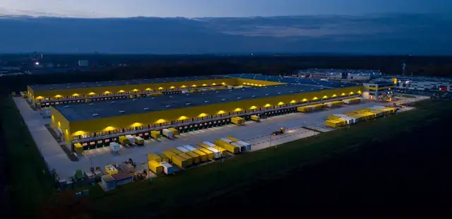 Aerial view of a massive, illuminated yellow distribution center at twilight with numerous trucks at the loading docks.