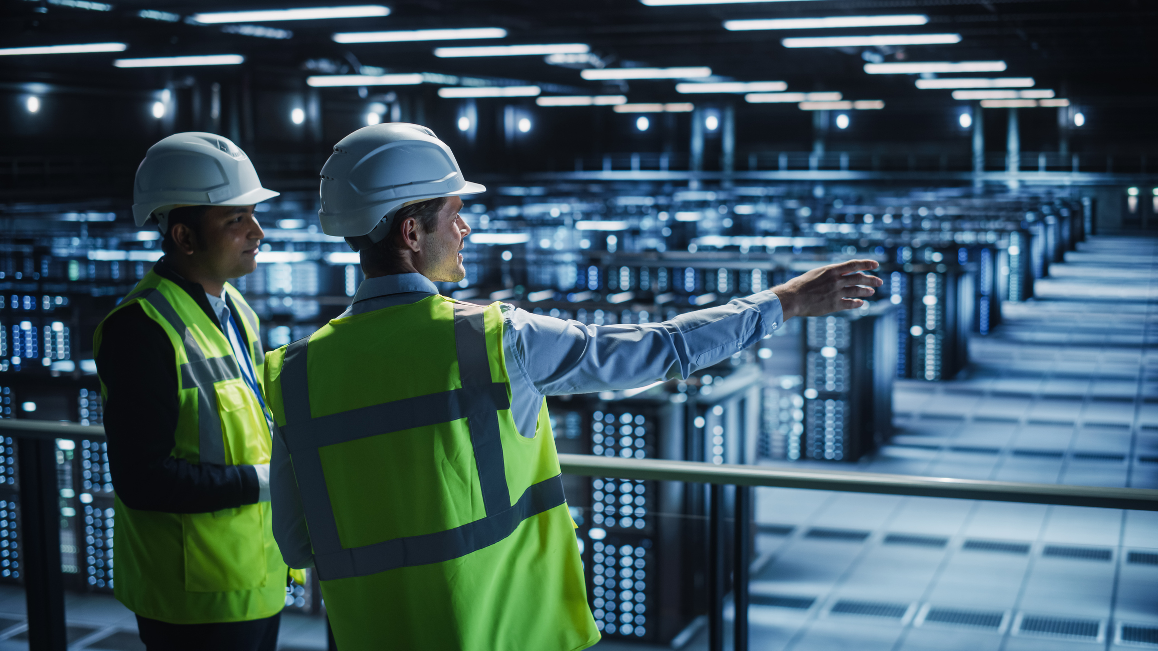 Two technicians in hard hats and safety vests overlooking rows of server racks from a catwalk